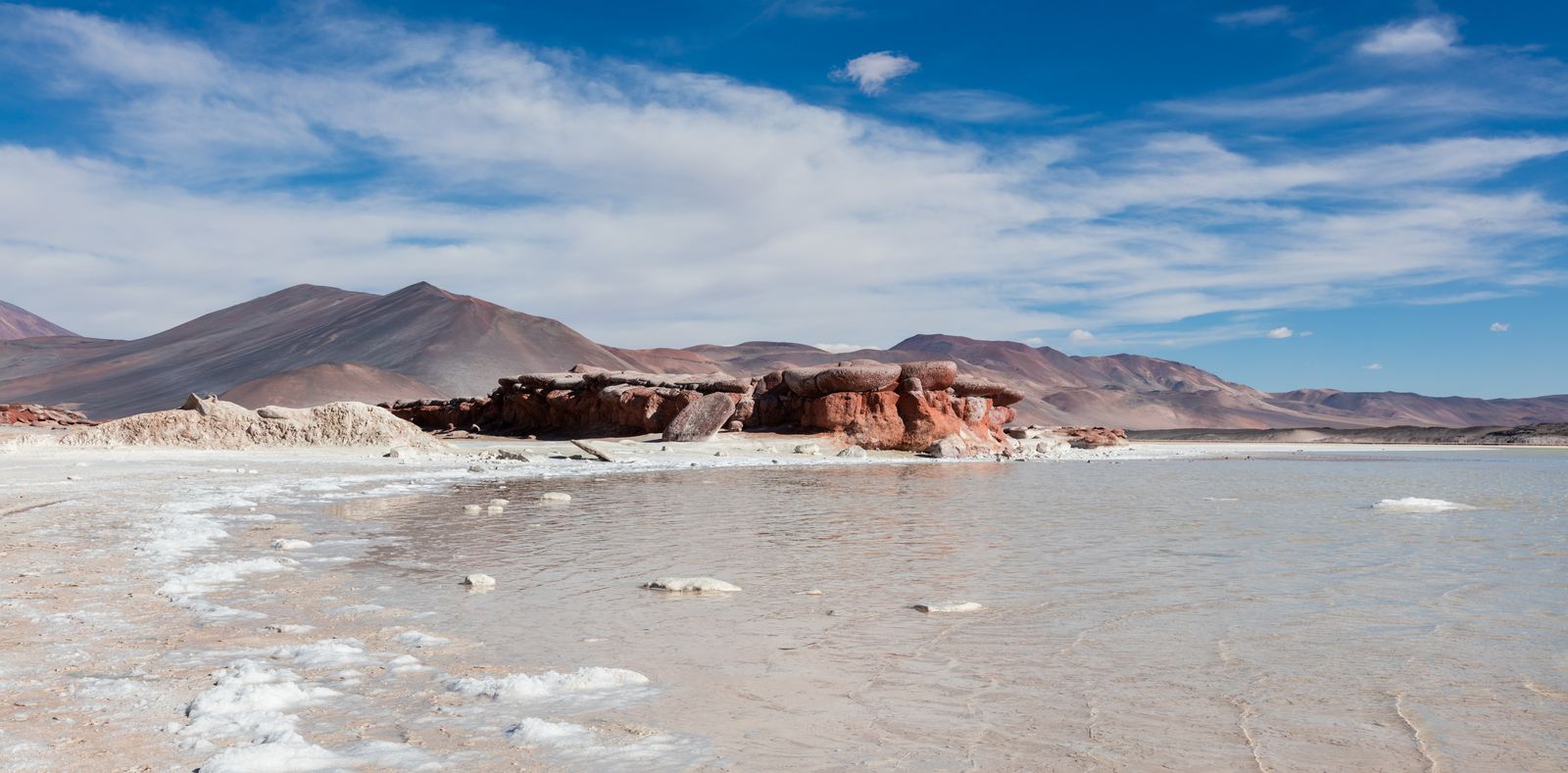 Red rocks beside the turquoise Aguas Calientes salt lake with volcanoes behind in northern Chile